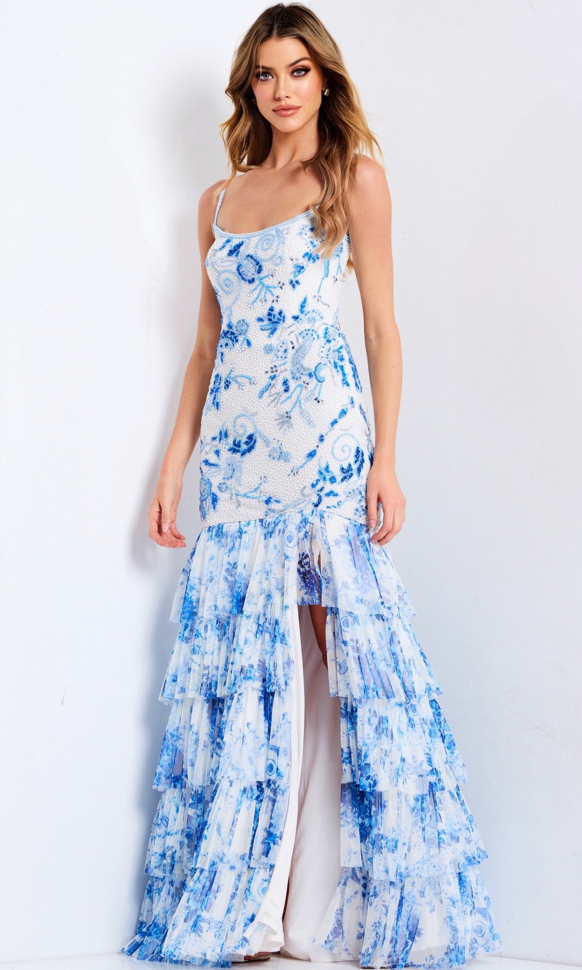 A woman in the Jovani 46873 Long Prom Dress, featuring a white and blue pattern, beaded floral embroidery, a fitted bodice, and a ruffle tiered skirt, stands against a plain light background.